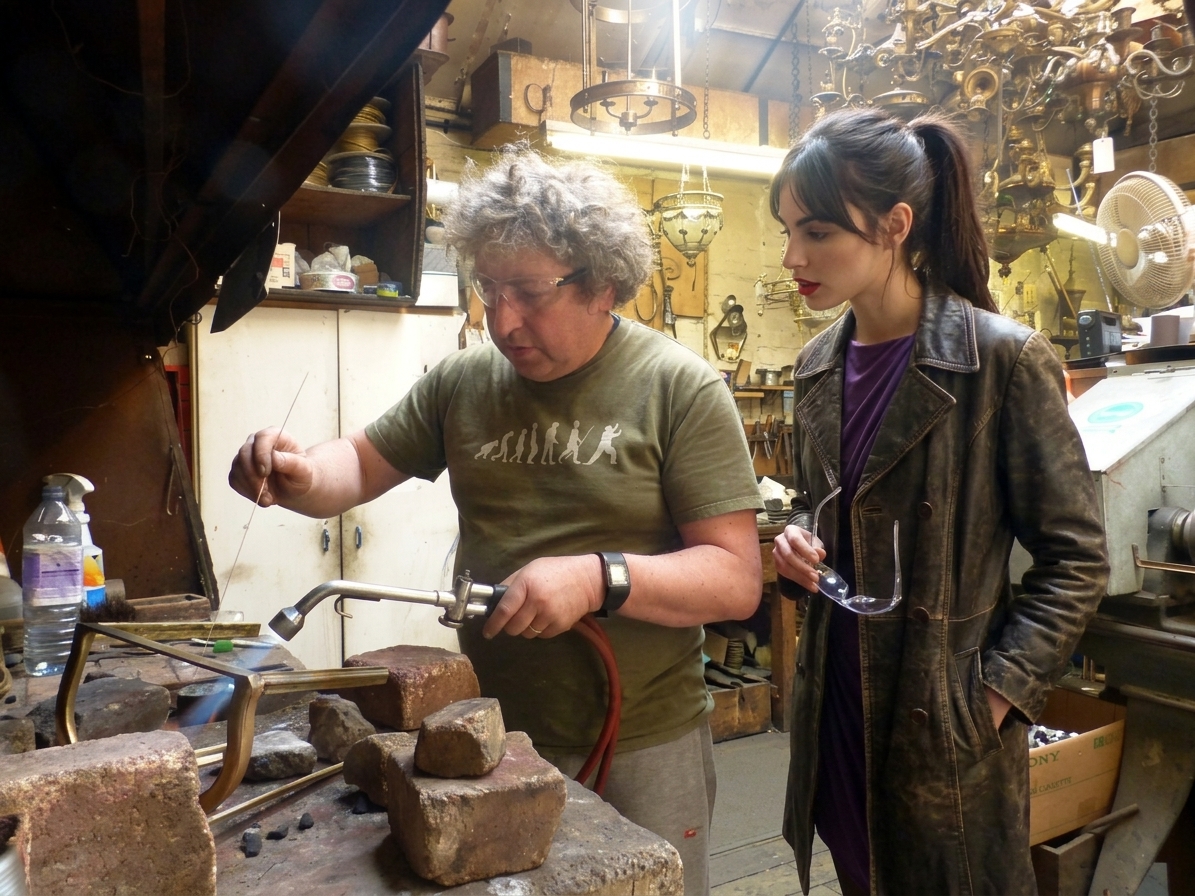 Cindy peering into the dusty workshop window, watching a focused workman at a bench assembling a brass electrical fixture